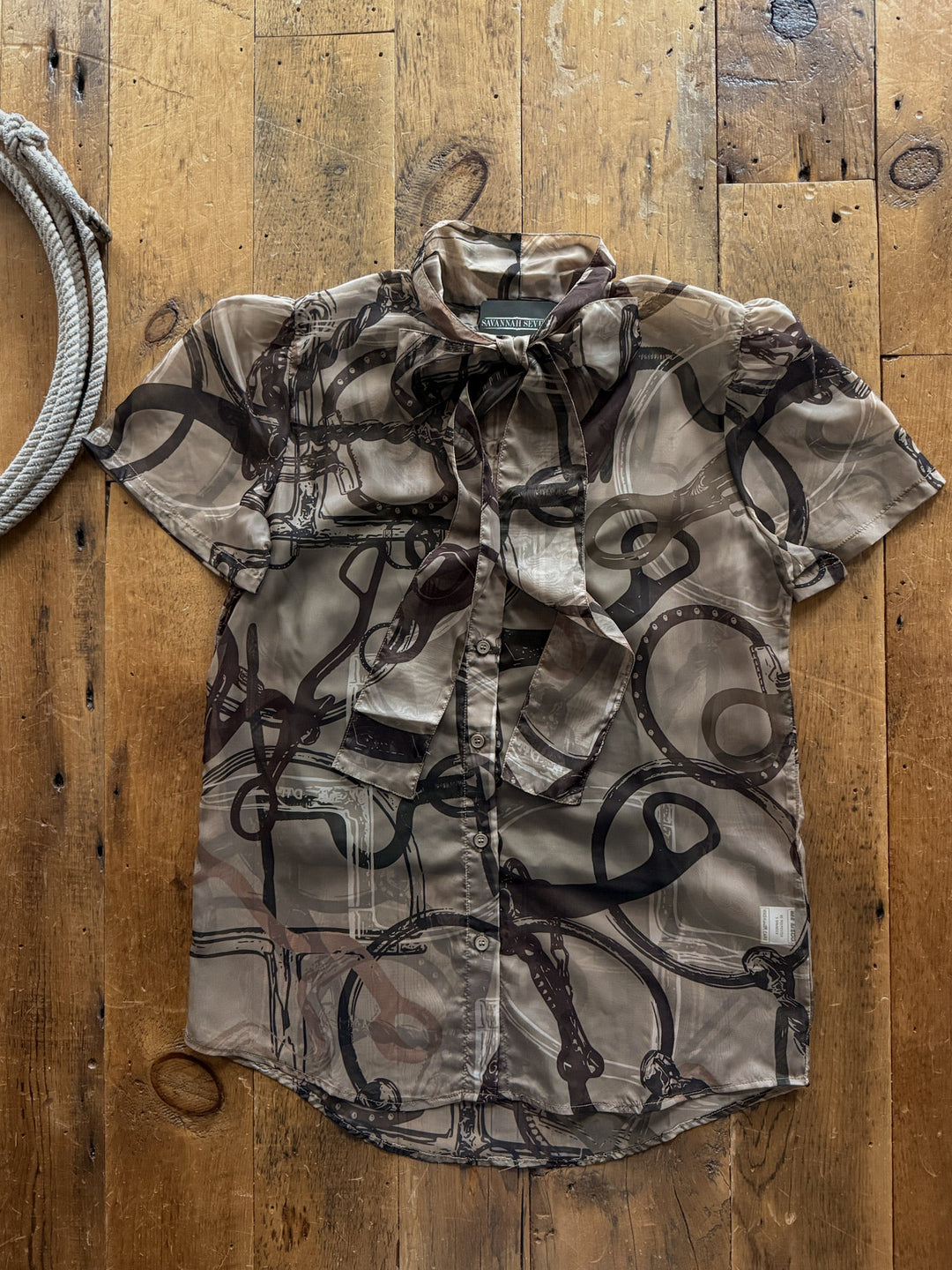 Bridle Stone Button Up Top featuring a relaxed-fit, short-sleeved design in a sheer tan fabric with a dark brown abstract bridle print and a tie-neck collar.