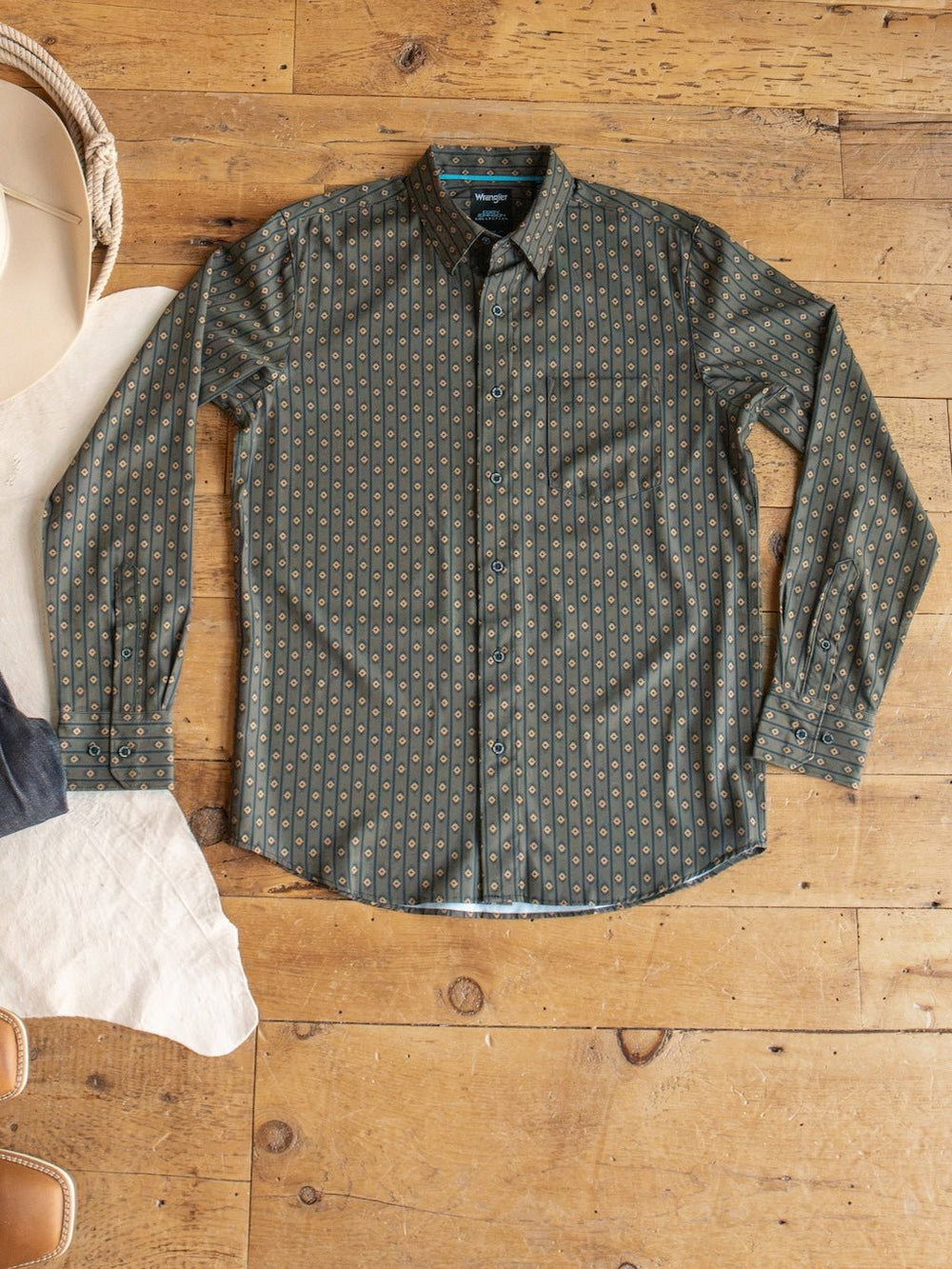 A Brigham Wrangler Men's Southwest Button Up shirt from Savannah Sevens western life{&}style, featuring a green color with a small, repeating pattern, is laid flat on a wooden floor. Part of a beige hat and tan shoes are visible at the edge of the image.
