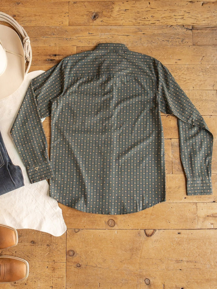 The Brigham Wrangler Men's Southwest Button Up by Savannah Sevens western life{&}style, a green long sleeve shirt with a moisture wicking design and classic button up style, is laid flat with the back side facing up on a wooden floor next to a hat, jeans, and brown boots.