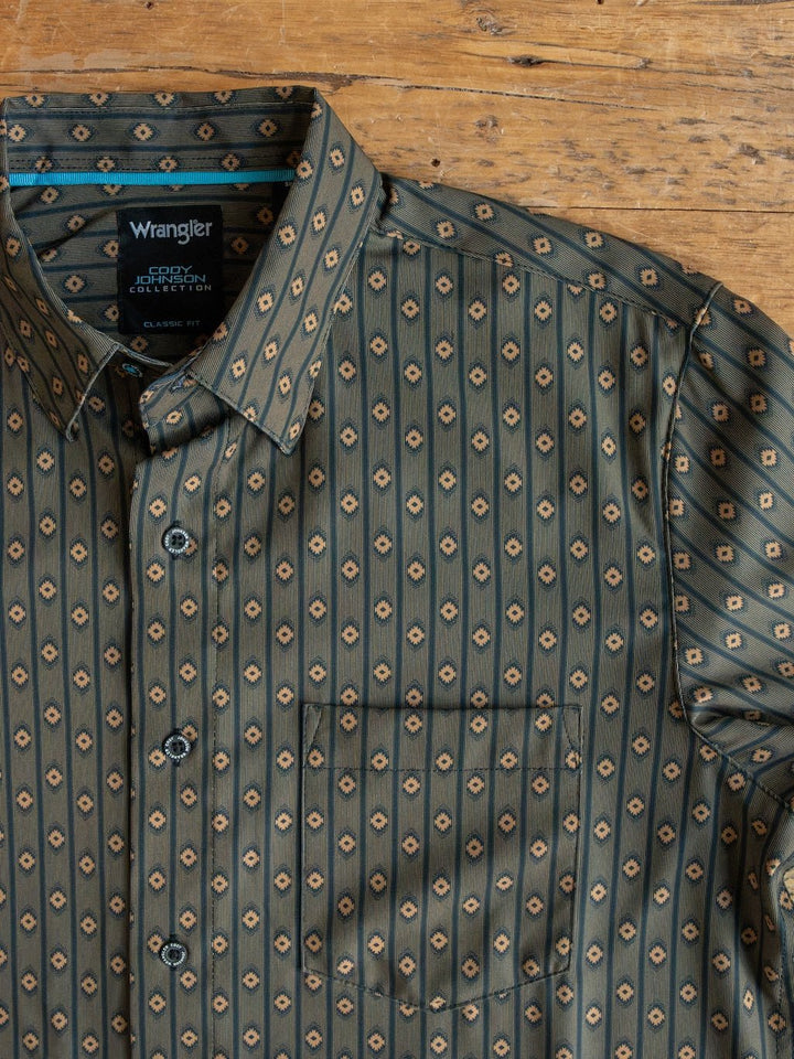 The Brigham Wrangler Men's Southwest Button Up from Savannah Sevens western life{&}style is shown here in green, featuring a repeating diamond design in tan and dark green. Laid flat on a wooden surface, the shirt displays its collar and left chest pocket.
