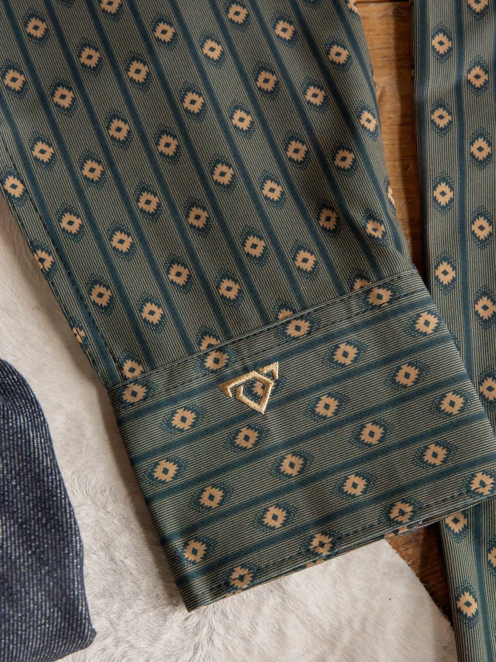 A green patterned sleeve from the Brigham Wrangler Men's Southwest Button Up by Savannah Sevens western life{&}style, featuring repeating geometric designs and a small gold triangular pin on the cuff, is shown lying on a light-colored textured surface next to another fabric.