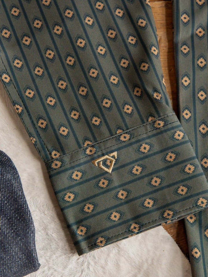 A green patterned sleeve from the Brigham Wrangler Men's Southwest Button Up by Savannah Sevens western life{&}style, featuring repeating geometric designs and a small gold triangular pin on the cuff, is shown lying on a light-colored textured surface next to another fabric.