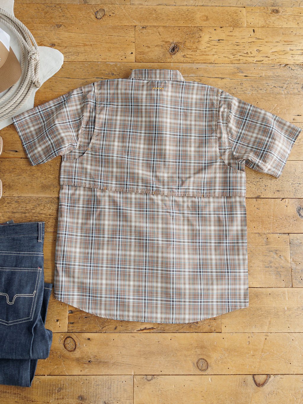 The Brimson Ariat Men's Plaid Button Up from Savannah Sevens western life{&}style in beige, brown, and blue is laid flat on a wooden floor with a straw hat and folded jeans nearby, blending style with moisture-wicking comfort.