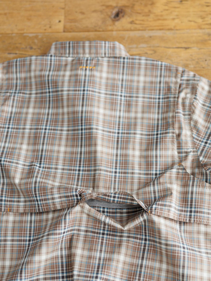 The Brimson Ariat Men's Plaid Button Up by Savannah Sevens western life{&}style, in brown, blue, and white plaid, is shown lying flat on a wooden surface with a visible tear below the back shoulder.