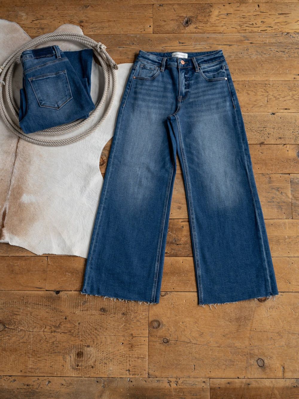 The Britton High Rise Wide Leg Crop Jean by Savannah Sevens western life{&}style lies flat on a wooden floor next to a folded jean, coiled rope, and cowhide rug.