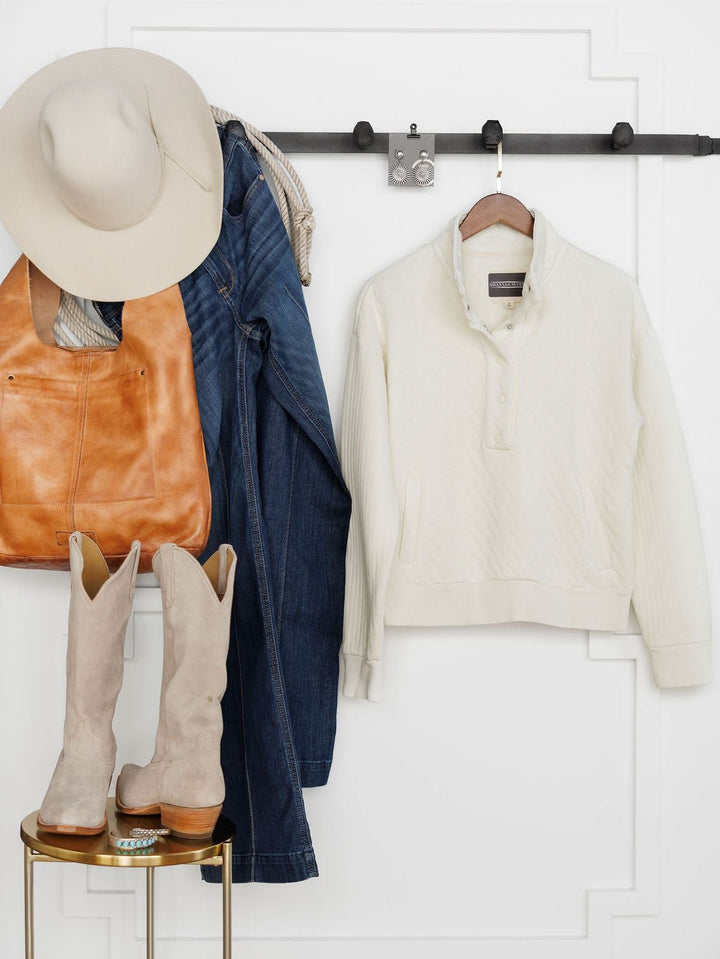 Clothing and accessories hang on a black rack: a beige hat, tan leather bag, blue jeans, and the Savannah Sevens western life{&}style Britton Ivory Quilted Pullover with striped scarf. Light cowboy boots sit on a small round table below.