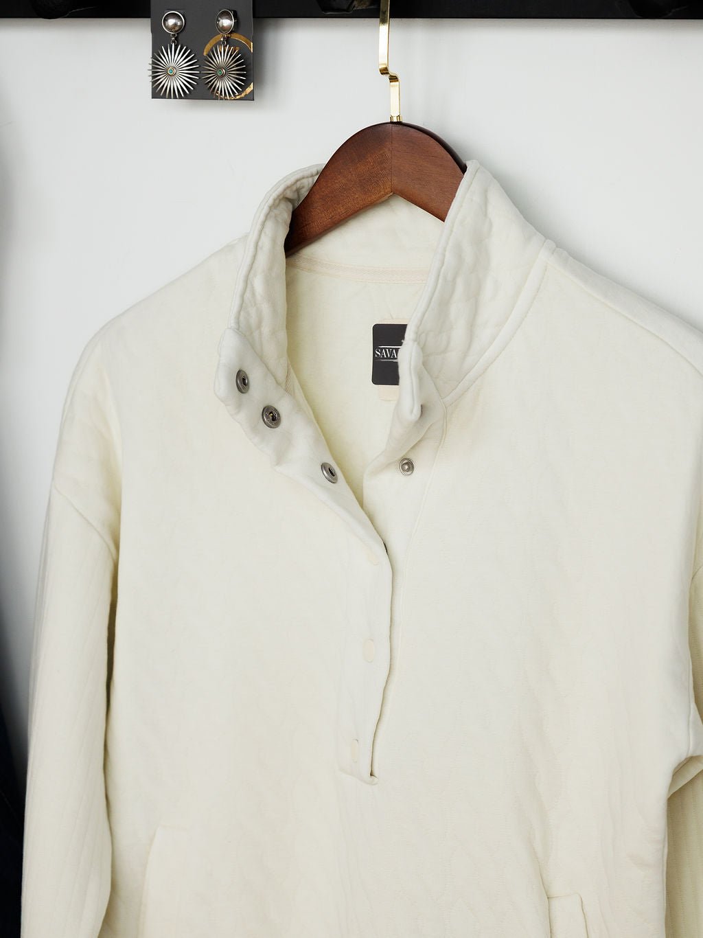 The Britton Ivory Quilted Pullover by Savannah Sevens western life{&}style, featuring long sleeves, a cream color, a three-silver-button collar, and a visible black neckline tag, hangs on a wooden hanger.