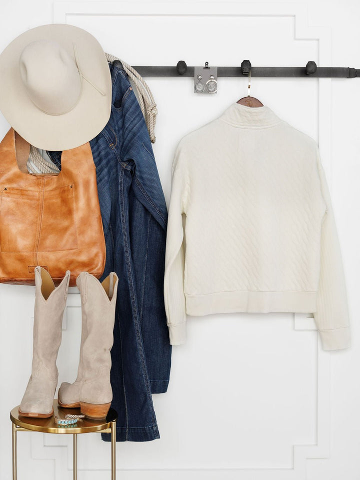 The Britton Ivory Quilted Pullover by Savannah Sevens western life{&}style hangs on a black wall rack with jeans, a beige hat, and tan bag above a table displaying tall beige suede boots, all set against a white paneled wall.