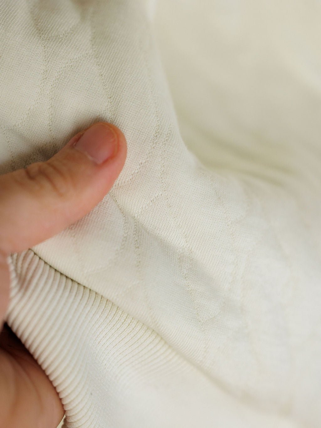 A close-up of a hand holding the Britton Ivory Quilted Pullover from Savannah Sevens western life{&}style, featuring off-white fabric with subtle diamond patterns and ribbed edging.