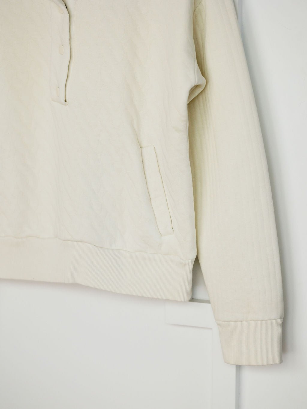 The Britton Ivory Quilted Pullover by Savannah Sevens western life{&}style features long sleeves, a textured quilted pattern, partial snap placket, side pocket, and ribbed cuffs and hem, shown hanging against a white background.