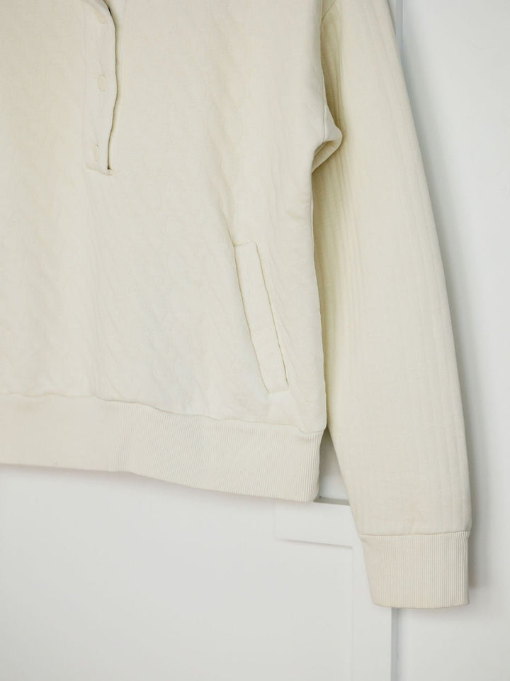 The Britton Ivory Quilted Pullover by Savannah Sevens western life{&}style features long sleeves, a textured quilted pattern, partial snap placket, side pocket, and ribbed cuffs and hem, shown hanging against a white background.