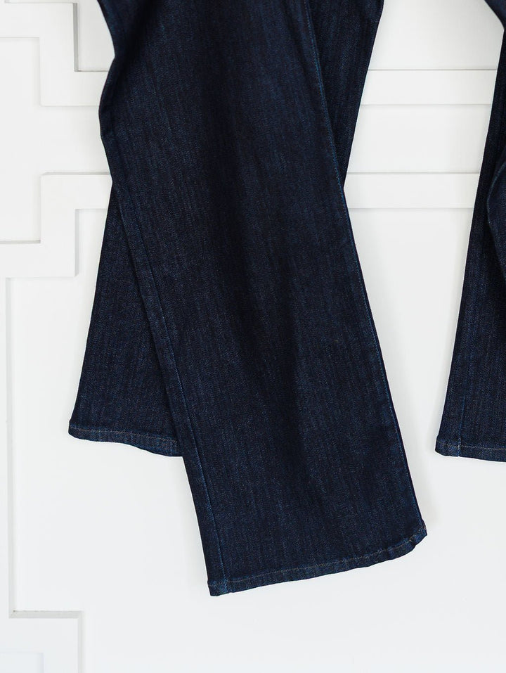 Close-up of The Brody High Rise Wide Leg Jean by Savannah Sevens western life{&}style, shown in dark wash denim with a slight flare, visible hem stitching, and wide leg cut against a white textured wall.