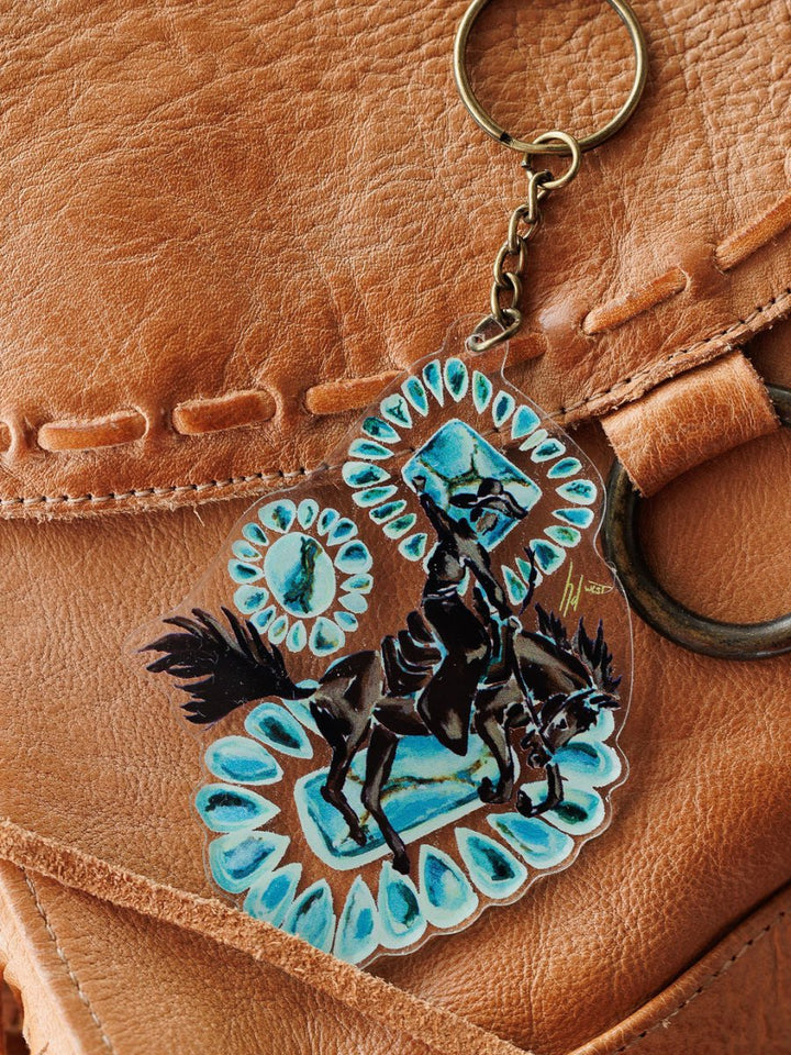 A close-up of the Bronc Keychain by Savannah Sevens western life{&}style, featuring horse and rider art with blue accents, shown on a brown leather bag with visible stitching and a metal ring.
