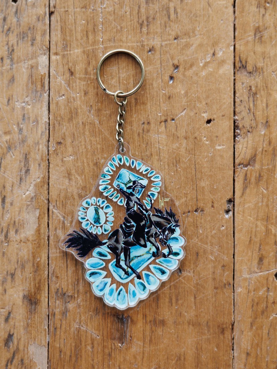 The Bronc Keychain from Savannah Sevens western life{&}style features a hand drawn rider on horseback with blue and white patterns, displayed on a worn wooden surface.