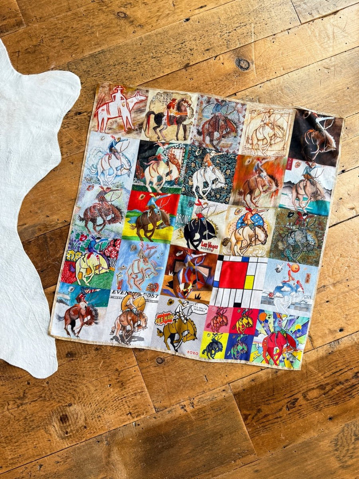 A Bronc Neck Scarf by Savannah Sevens western life{&}style, featuring colorful squares with retro bronc horse and rider graphics in various artistic styles, is displayed on a wooden floor beside a white cowhide rug.