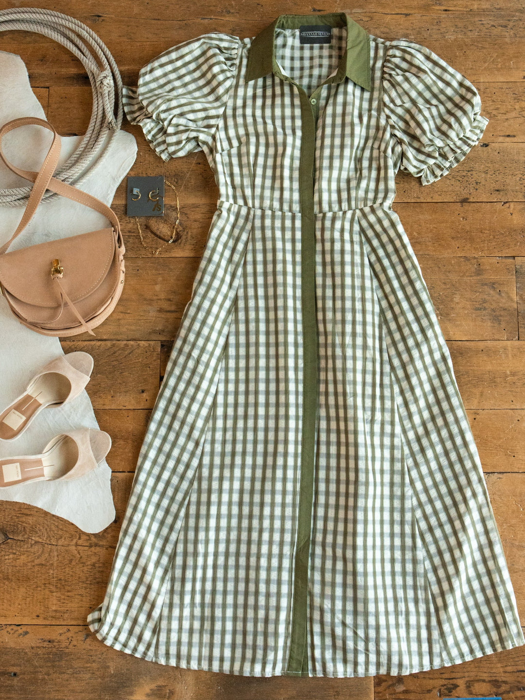 The Brookville Olive Gingham Puff Sleeve Dress by Savannah Sevens western life{&}style is laid out on a wooden floor, styled alongside beige heels and a tan purse.