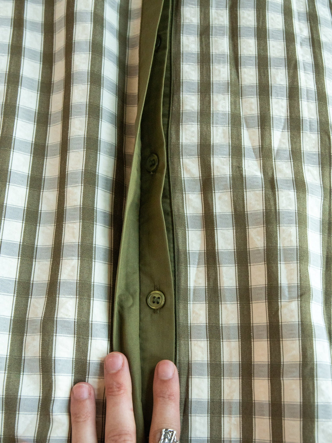 A hand adorned with a silver ring holds open the Brookville Olive Gingham Puff Sleeve Dress by Savannah Sevens western life{&}style, showcasing the inner green button placket with visible buttons.