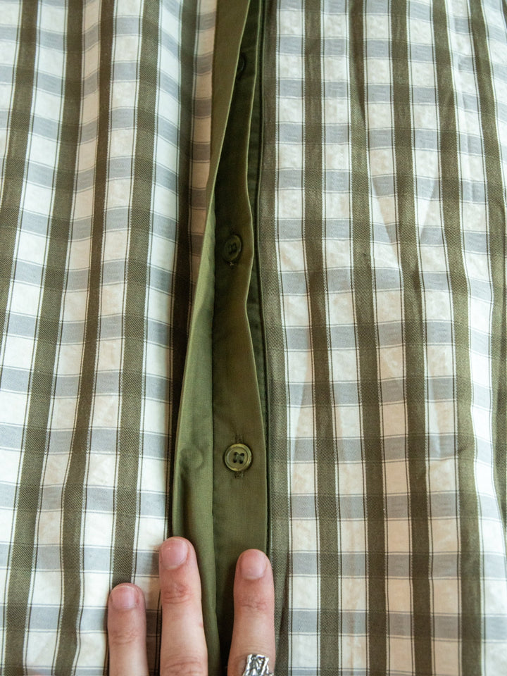 A hand adorned with a silver ring holds open the Brookville Olive Gingham Puff Sleeve Dress by Savannah Sevens western life{&}style, showcasing the inner green button placket with visible buttons.