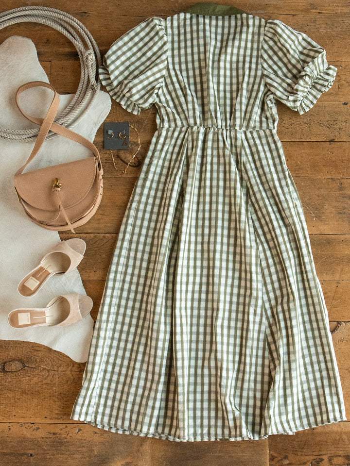 The Brookville Olive Gingham Puff Sleeve Dress by Savannah Sevens western life{&}style is displayed on a wooden floor alongside beige sandals, a tan crossbody bag, jewelry, a coiled rope, and a white cowhide rug.