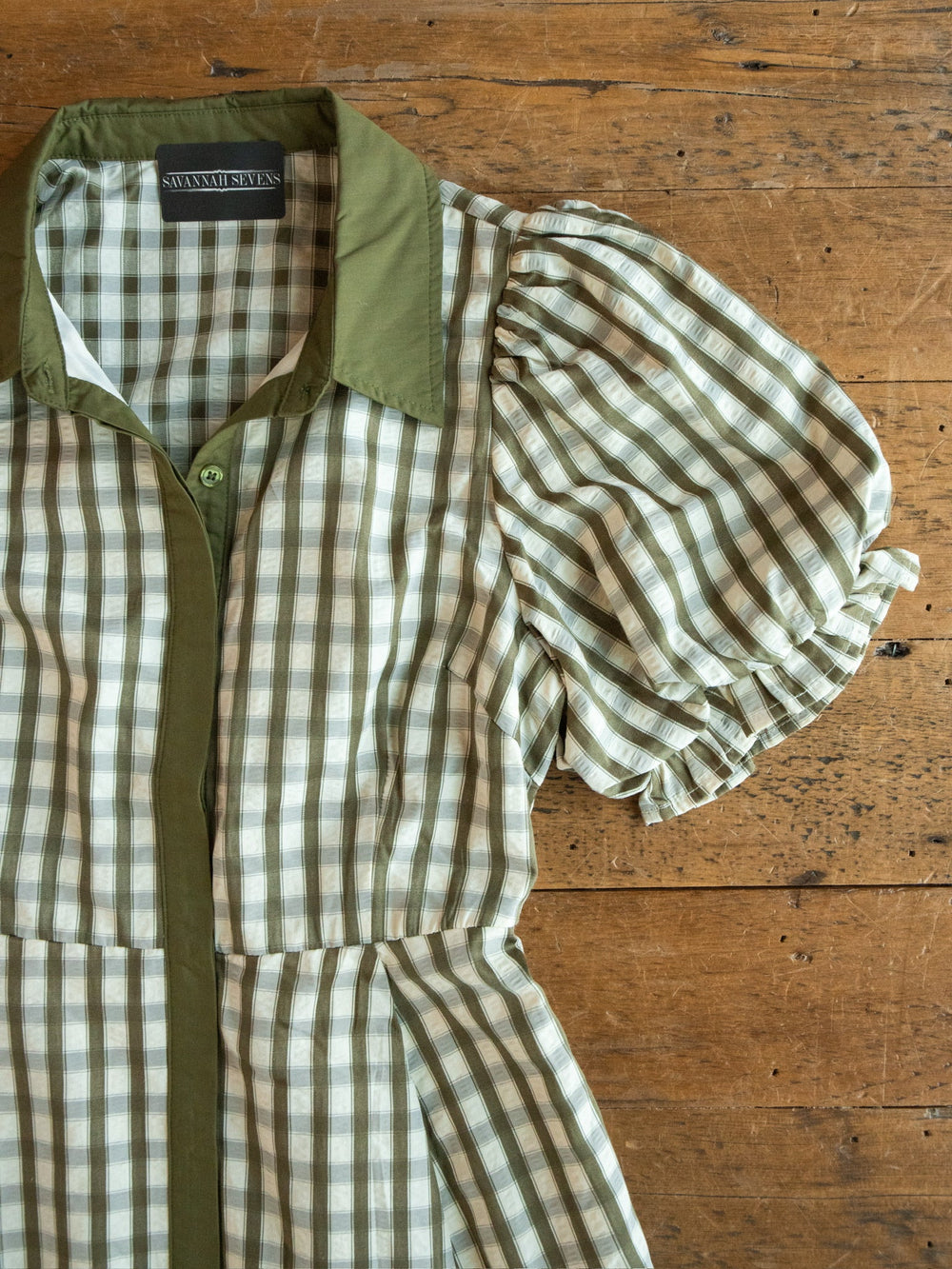 Brookville Olive Gingham Puff Sleeve Dress by Savannah Sevens western life{&}style, featuring a green collar, is laid flat on a wooden surface.