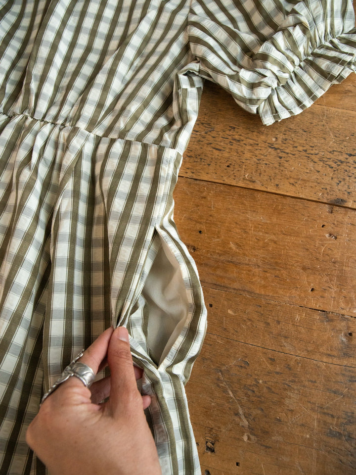 A hand adorned with silver rings displays the pocket of the Brookville Olive Gingham Puff Sleeve Dress by Savannah Sevens western life{&}style, laid out on a wooden surface.