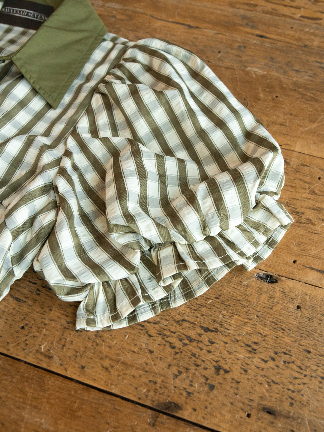 The Brookville Olive Gingham Puff Sleeve Dress by Savannah Sevens western life{&}style, featuring green and white gingham with puff sleeves and ruffled cuffs, is laid out on a worn wooden table.