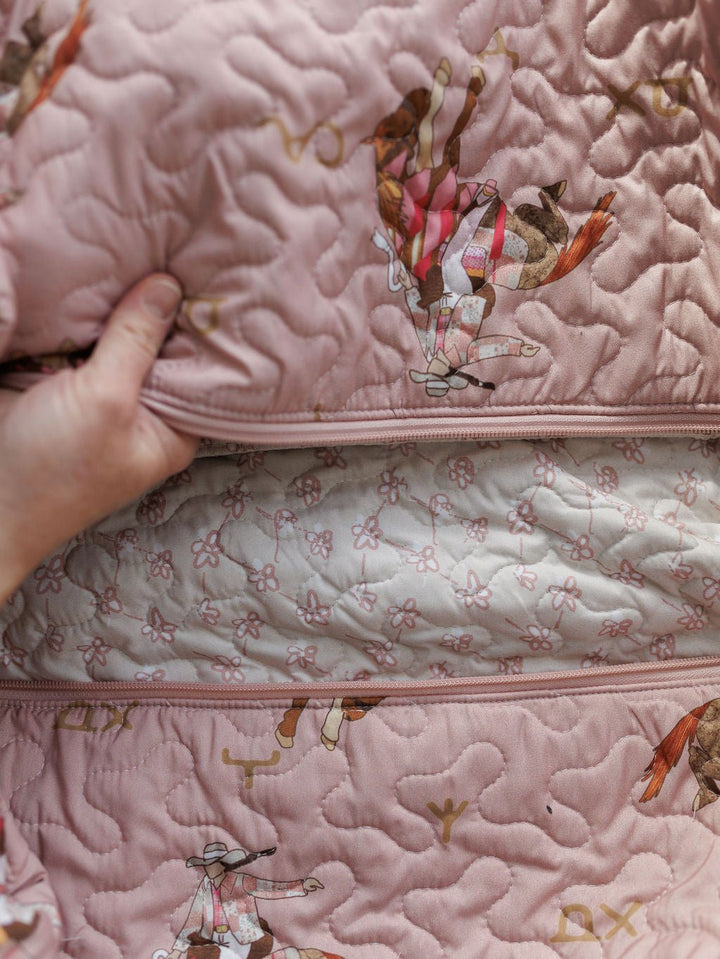 A hand pulls back Savannah Sevens western life{&}style's Brumby Rose Collection pink quilted fabric with cowboy and horse motifs, revealing a light pink floral-patterned inner lining.