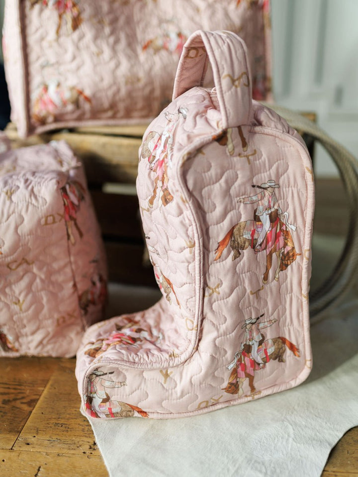 A pink quilted boot bag from the Brumby Rose Collection by Savannah Sevens western life{&}style, featuring red and brown horse rider patterns, sits on a wooden surface beside matching bags in the same design.