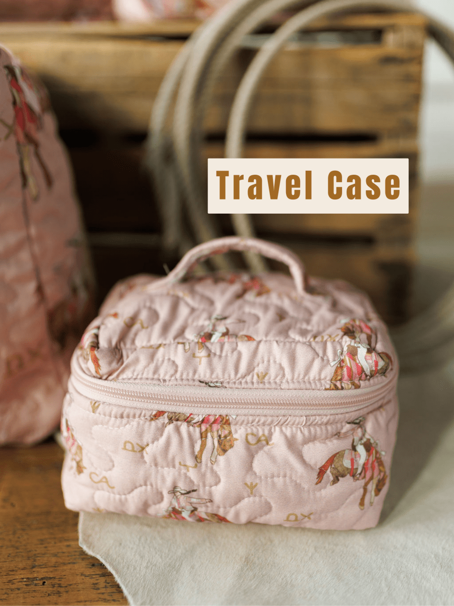 The Brumby Rose Collection travel case by Savannah Sevens western life&style, featuring a pink quilted horse pattern, sits on a wooden surface before a rustic crate with "Travel Case" text above.