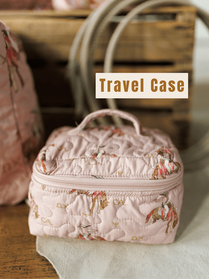 The Brumby Rose Collection travel case by Savannah Sevens western life&style, featuring a pink quilted horse pattern, sits on a wooden surface before a rustic crate with "Travel Case" text above.