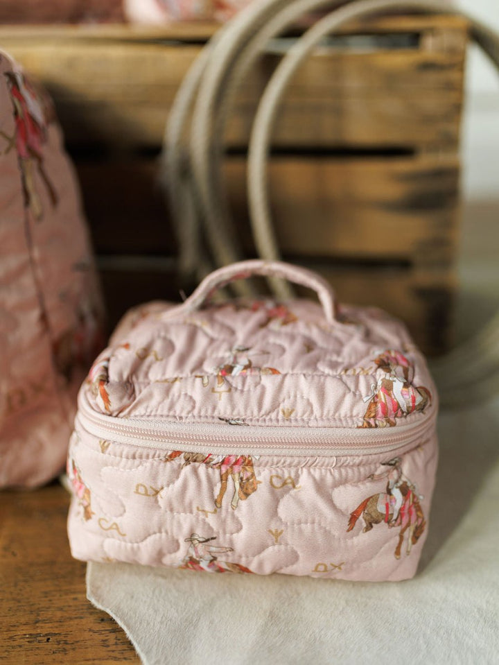 A small, quilted pink Brumby Rose bag from Savannah Sevens western life{&}style sits on a wooden surface in front of a crate and coiled rope, featuring a horse and rider print.