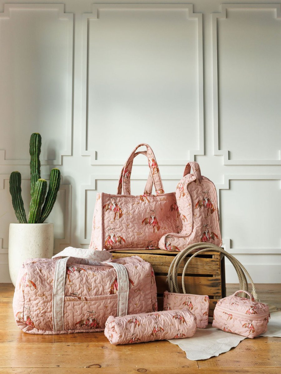 The Brumby Rose Collection by Savannah Sevens western life&style features pink quilted floral bags—duffels, backpack, and pouches—styled on wooden crates and a rug with a tall potted cactus before a white paneled wall.