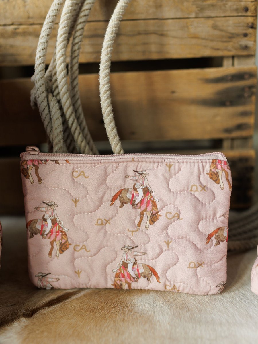 A pink quilted pouch from the Brumby Rose Collection by Savannah Sevens western life&style, featuring rodeo cowgirl illustrations on horseback with rope and crate details, displayed on a light brown animal hide.