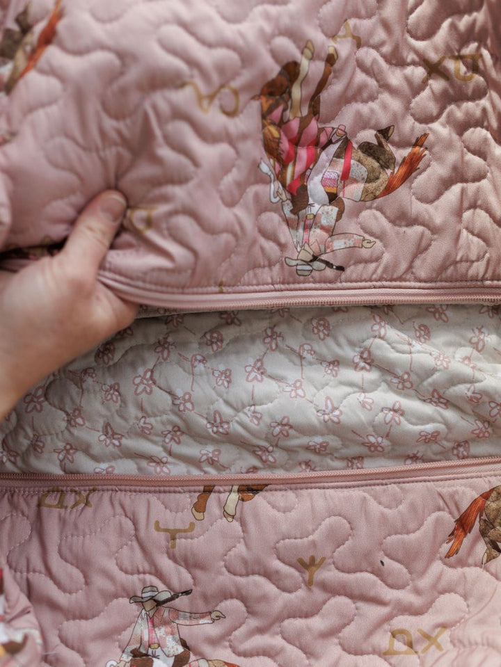 A hand pulls back Savannah Sevens western life{&}style's Brumby Rose Collection pink quilted fabric with cowboy and horse motifs, revealing a light pink floral-patterned inner lining.