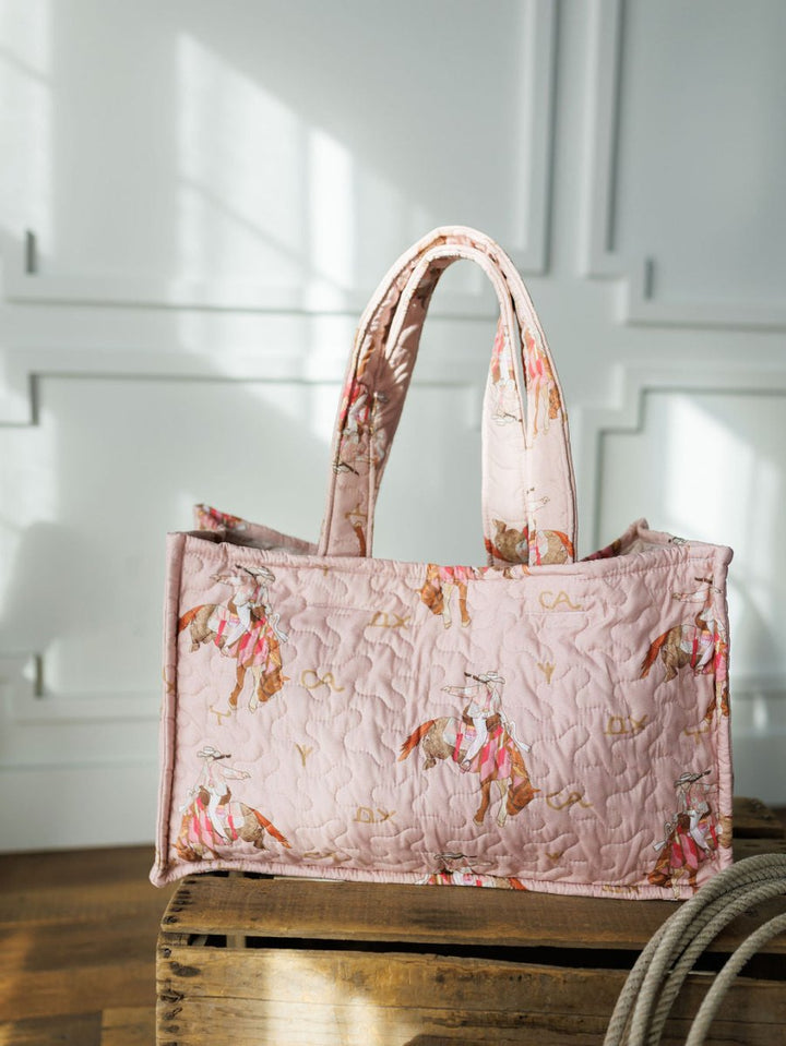 The Brumby Rose tote from Savannah Sevens western life{&}style, featuring quilted pink fabric and sturdy handles, rests on a wooden crate in a sunlit room with white paneled walls.