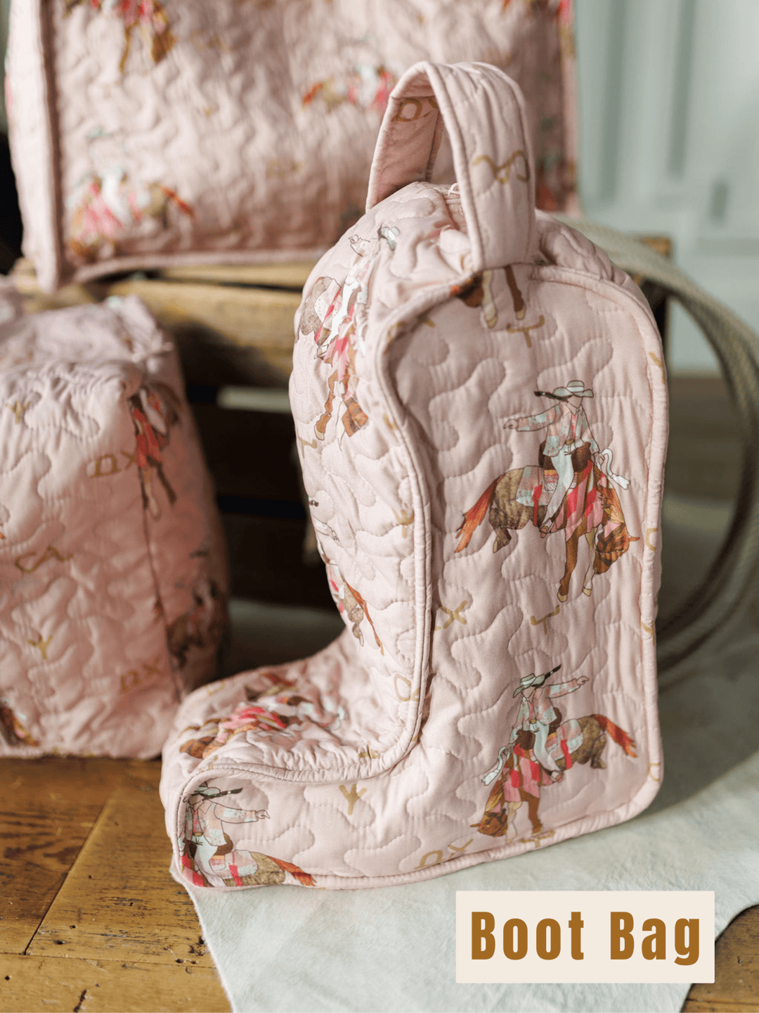 A pink quilted boot-shaped bag featuring horses and riders from the Brumby Rose Collection by Savannah Sevens western life{&}style sits on a wooden surface near similar quilted items, with a "Boot Bag" label in the corner.