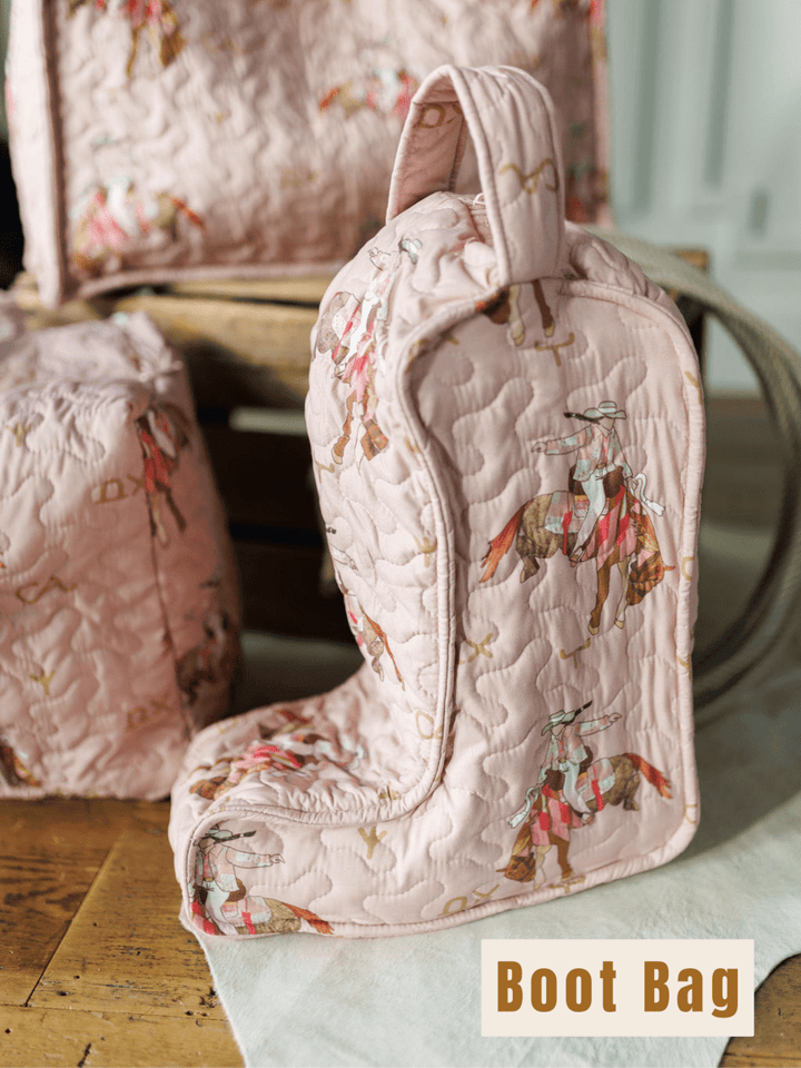 A pink quilted boot-shaped bag featuring horses and riders from the Brumby Rose Collection by Savannah Sevens western life{&}style sits on a wooden surface near similar quilted items, with a "Boot Bag" label in the corner.