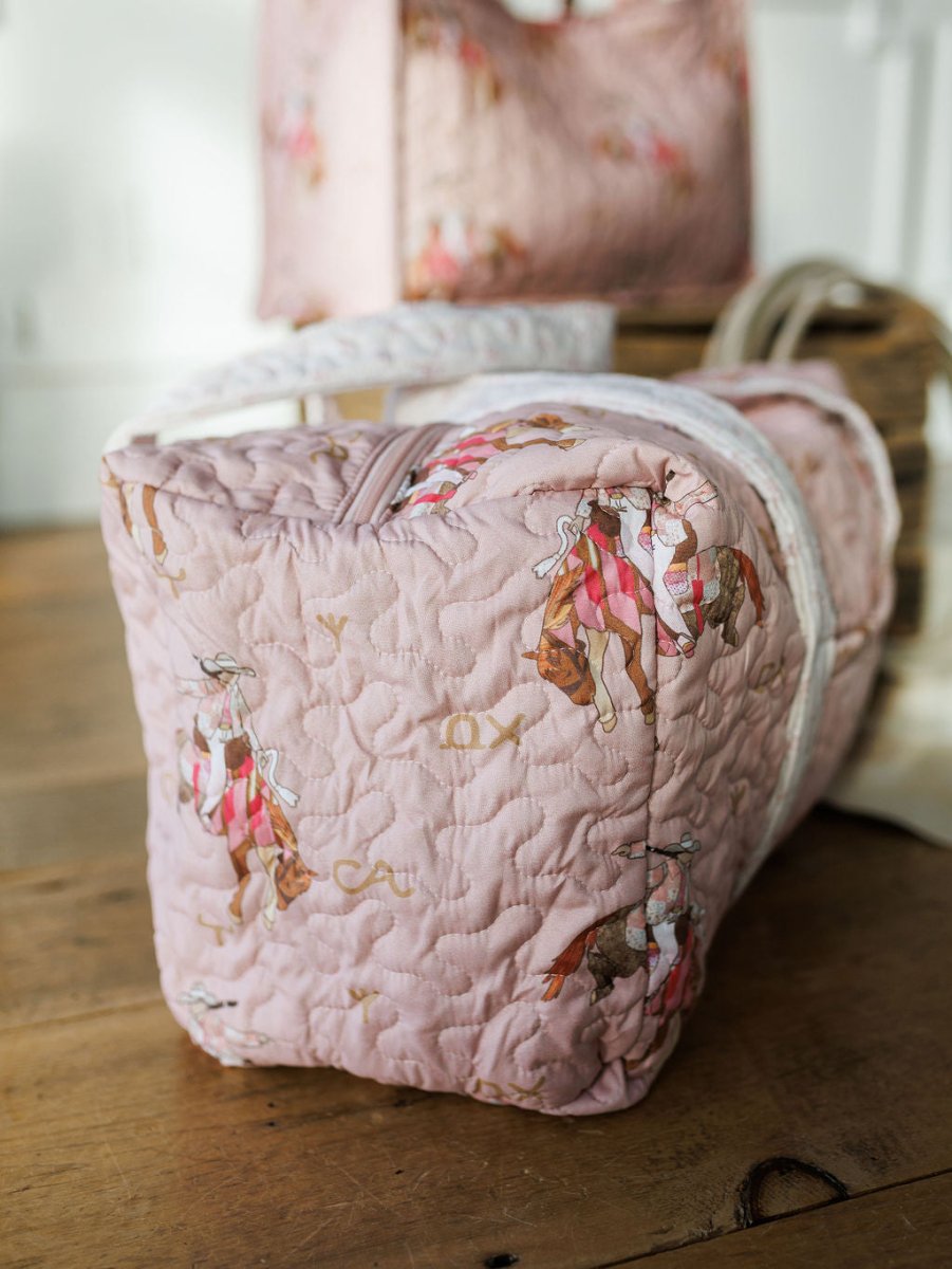 The Brumby Rose quilted pink duffel bag by Savannah Sevens western life&style features a cowgirl hat and boot pattern, white handles, and zipper. Part of a quilted bag set from the western travel collection with a matching bag shown in back.