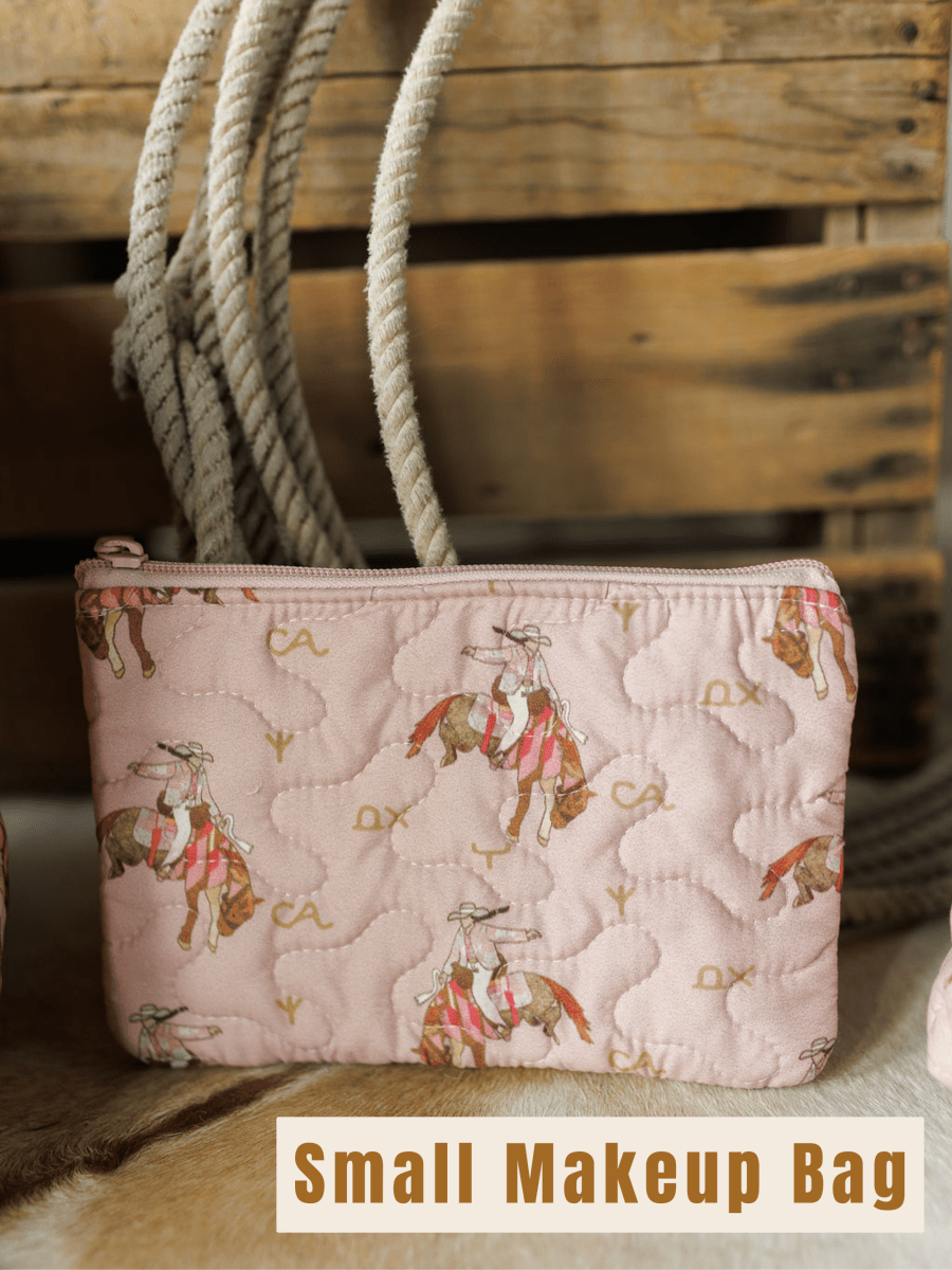 A small, quilted pink makeup bag with a cowboy and horse design from Savannah Sevens western life{&}style’s Brumby Rose {Collection}, displayed in front of coiled rope and a wooden crate. "Small Makeup Bag" is written below in bold letters.