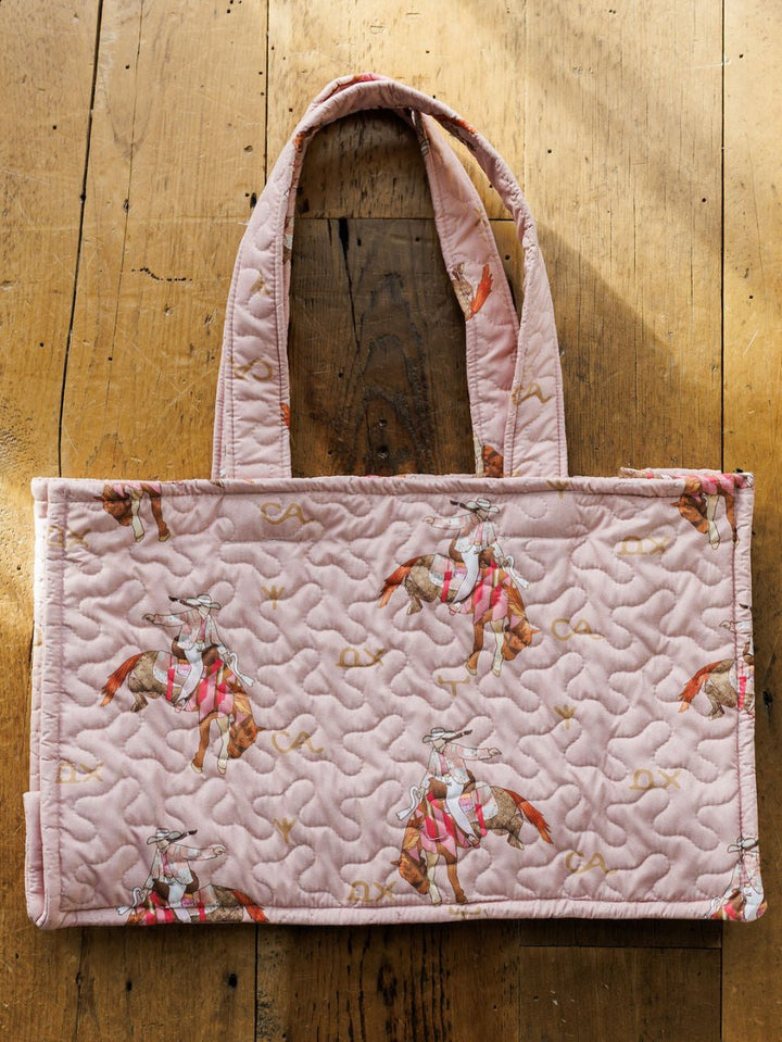 The Brumby Rose Collection by Savannah Sevens western life&style showcases a pink quilted bag set with cowboy and horse patterns, displayed on rustic wood in sunlight—a charming pick for western travel.