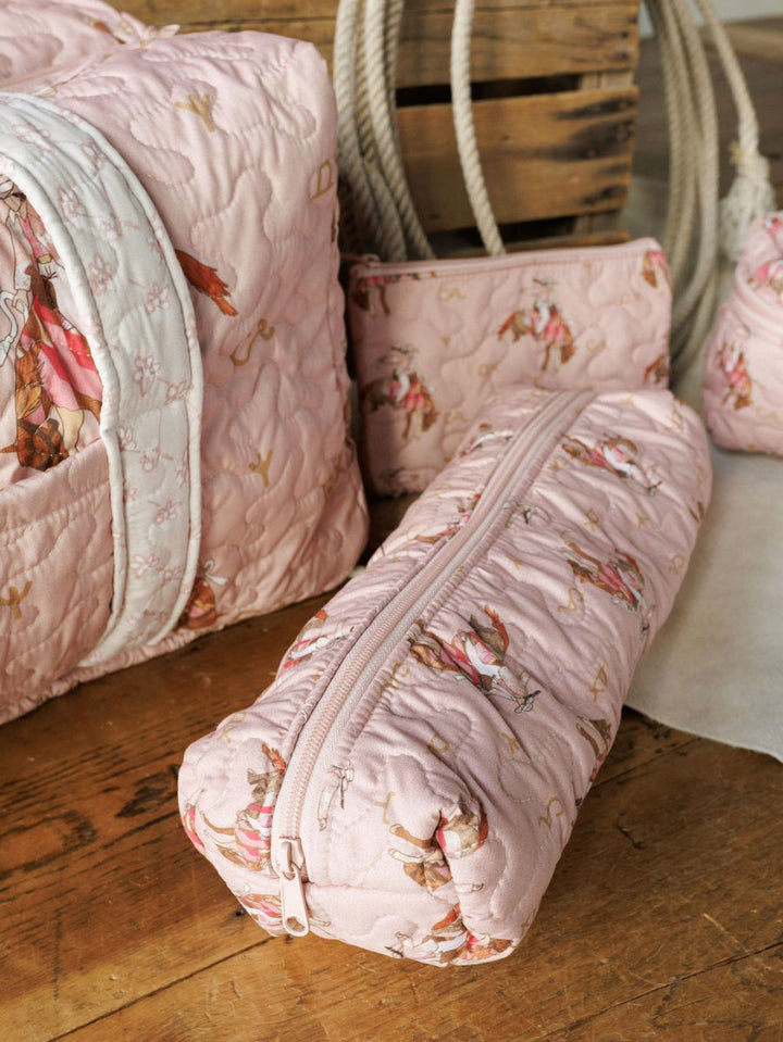 The Brumby Rose Collection by Savannah Sevens western life{&}style features pink quilted bags with whimsical illustrations and white zippers, styled on wood with crate and rope accents for a charming western travel vibe.