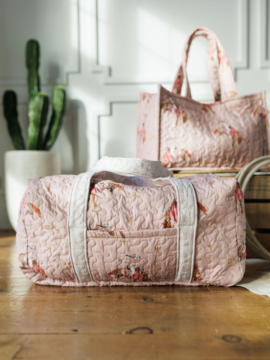 The Brumby Rose Collection from Savannah Sevens western life&style features a pink quilted floral bag set on a wooden floor in sunlight, paired with a matching tote and potted cactus—ideal for your western travel adventures.