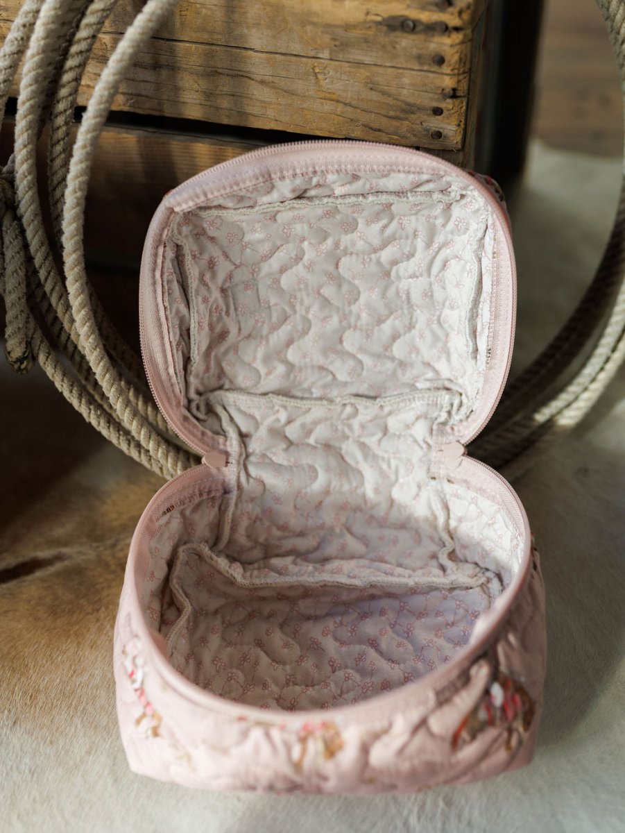 The Brumby Rose pouch from Savannah Sevens western life&style’s collection rests on a light surface. Quilted pink with a soft, padded interior, it’s empty; a coiled rope and wooden box are visible in the background.