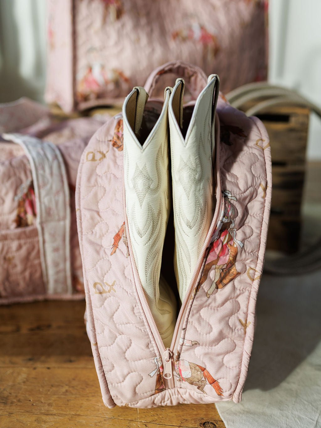 A Brumby Rose quilted pink bag from Savannah Sevens western life & style is open on a wooden surface, displaying white cowboy boots inside. Its horse-and-rider pattern stands out while similar bags blur in the background.