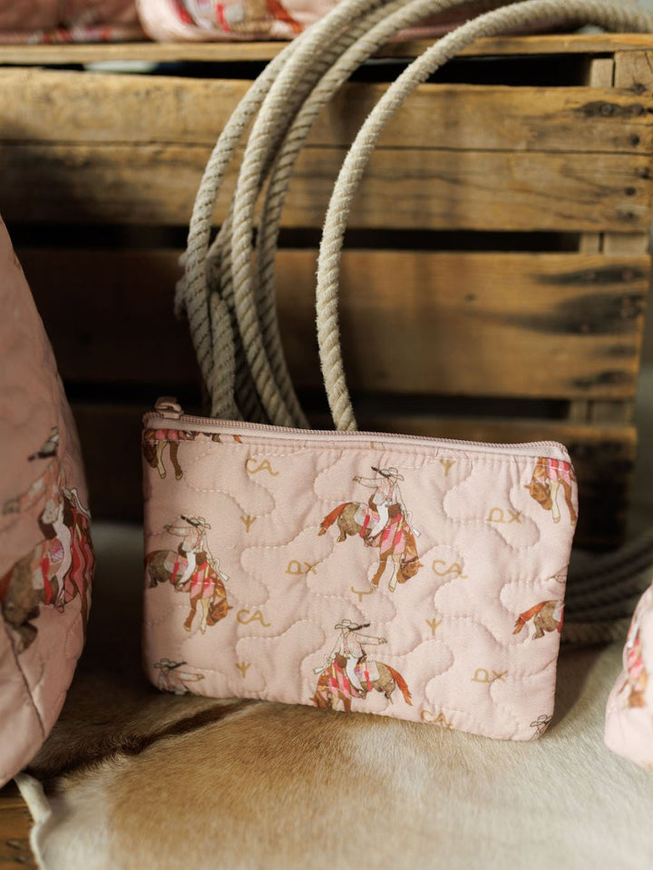 The Brumby Rose Collection quilted pink pouch by Savannah Sevens western life{&}style, with a rodeo cowboy and horse pattern, sits on animal hide beside a coiled rope and wooden crate.
