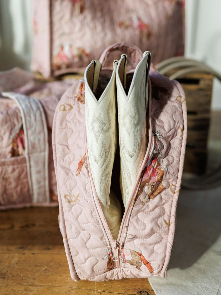 A Brumby Rose quilted pink bag from Savannah Sevens western life & style is open on a wooden surface, displaying white cowboy boots inside. Its horse-and-rider pattern stands out while similar bags blur in the background.