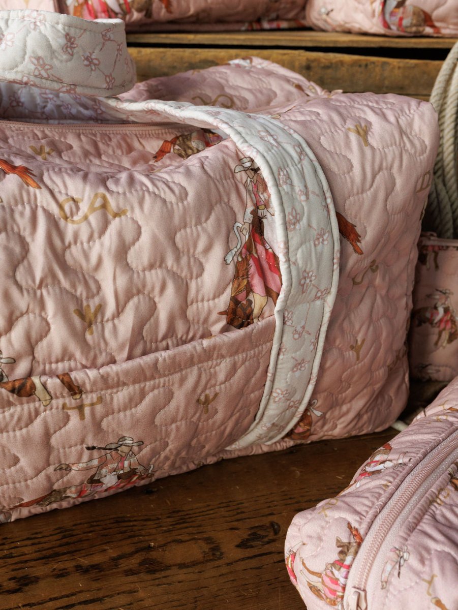 A close-up of the Brumby Rose collection bag by Savannah Sevens western life&style—soft, quilted pink fabric with white handles, zipper, and horse-and-rider print—rests on wood beside matching quilted pieces.