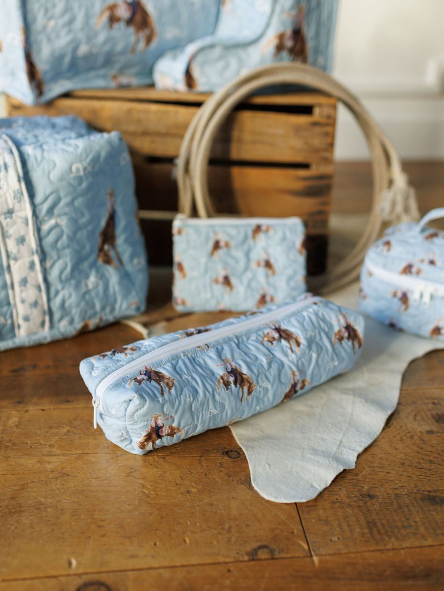 The Brumby Sky Collection by Savannah Sevens western life&style features a light blue quilted bag set with cowboy and horse print, displayed on wood beside a coiled rope, crate, and cowhide rug—ideal for any rodeo cowgirl or western traveler. By Ida Mae.