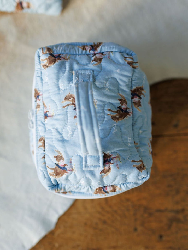The Brumby Sky Collection by Savannah Sevens western life{&}style features a light blue quilted bag set with a top handle and small horse and rider motifs, channeling a rodeo cowgirl vibe on wood and white cloth. By Ida Mae. 