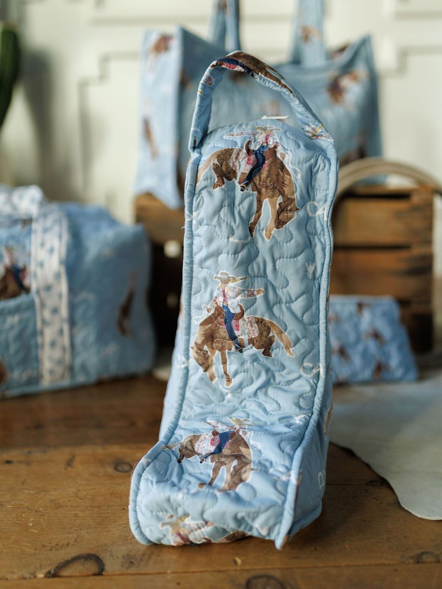 The Brumby Sky Collection bag by Savannah Sevens western life&style features a light blue quilted design with a cowboy and horse print, shown standing on wood with matching bags and crates blurred in the background. By Ida Mae.